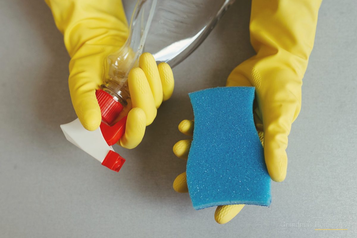 Yellow rubber gloves holding a sponge and spray bottle ready to scrub the dishwasher filter
